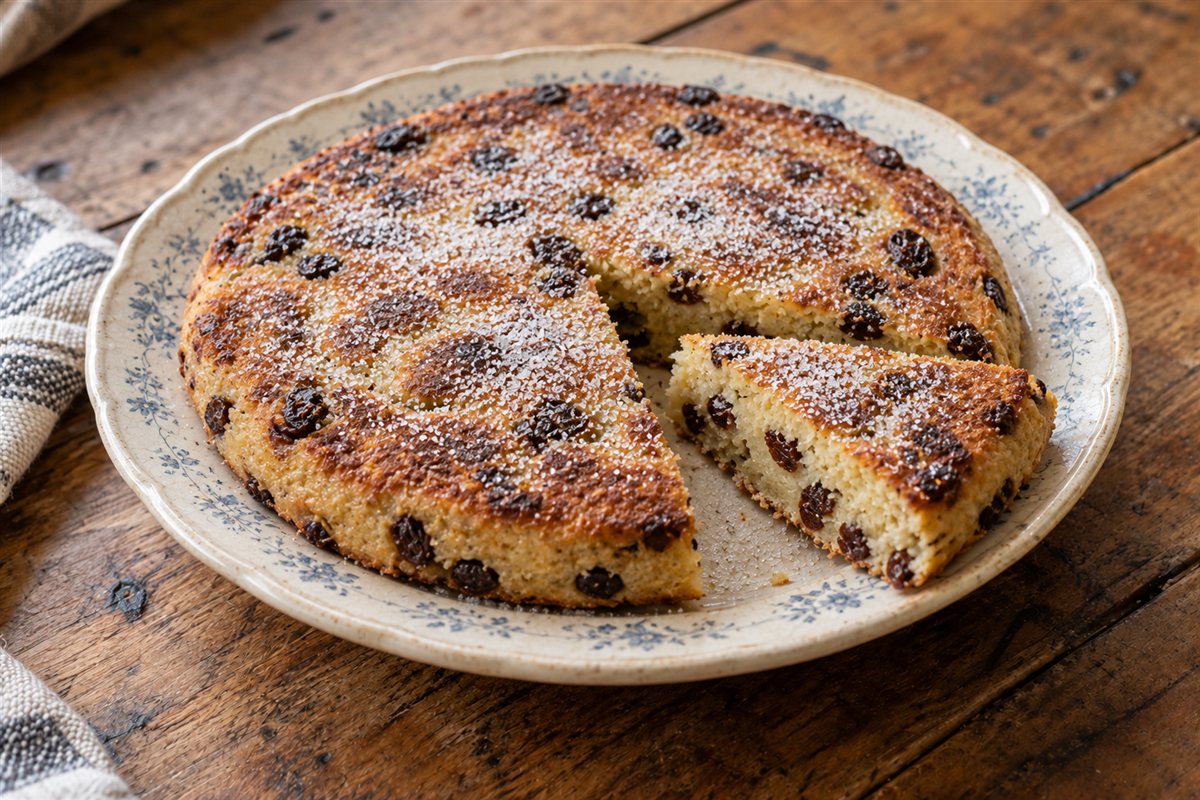 A flat golden fruit cake dusted with sugar on an old plate with one slice cut out